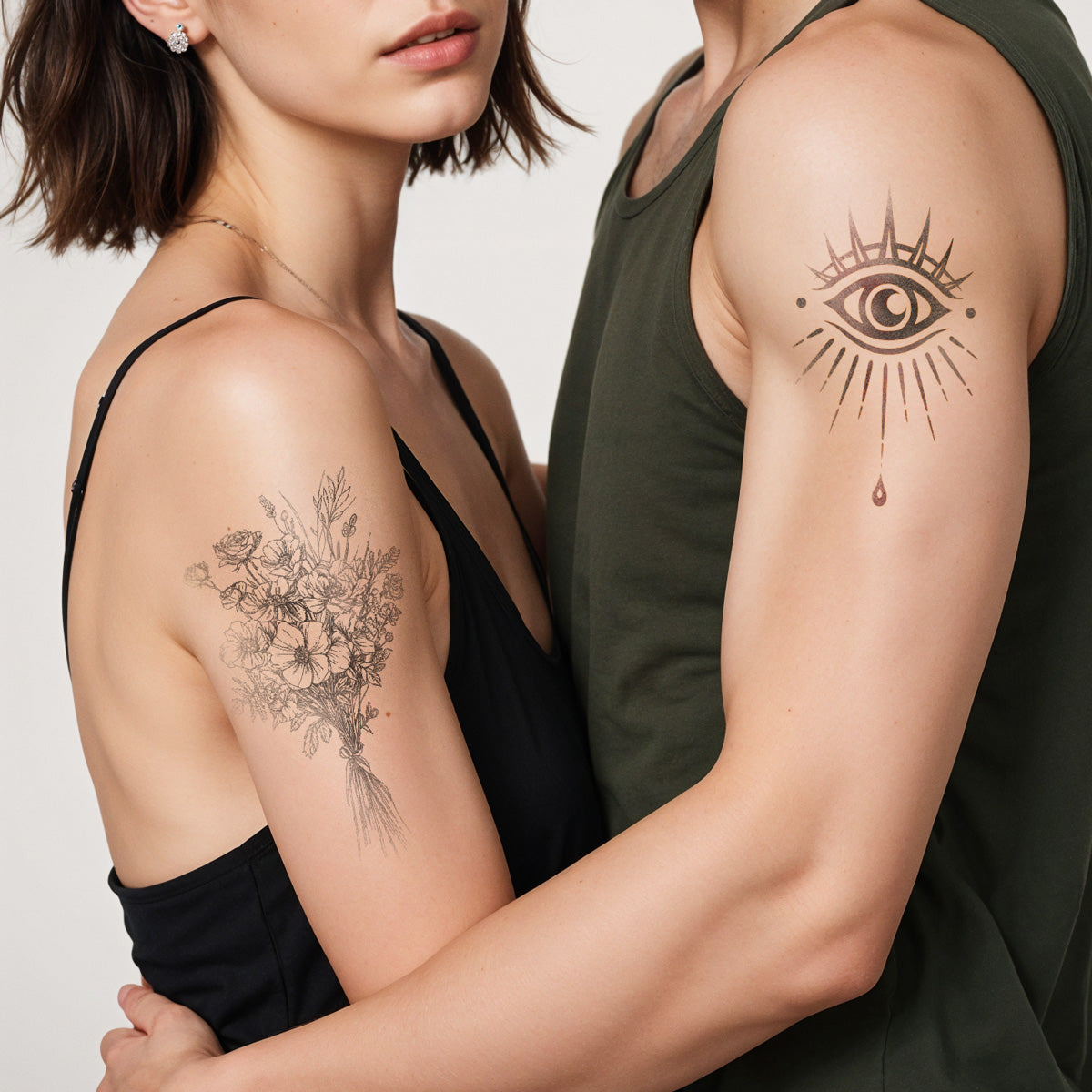 A couple with visible arm tattoos embraces. The woman has a detailed floral tattoo on her upper arm and wears a black top. The man has an eye tattoo with radiating lines and a teardrop on his upper arm and wears a green tank top.